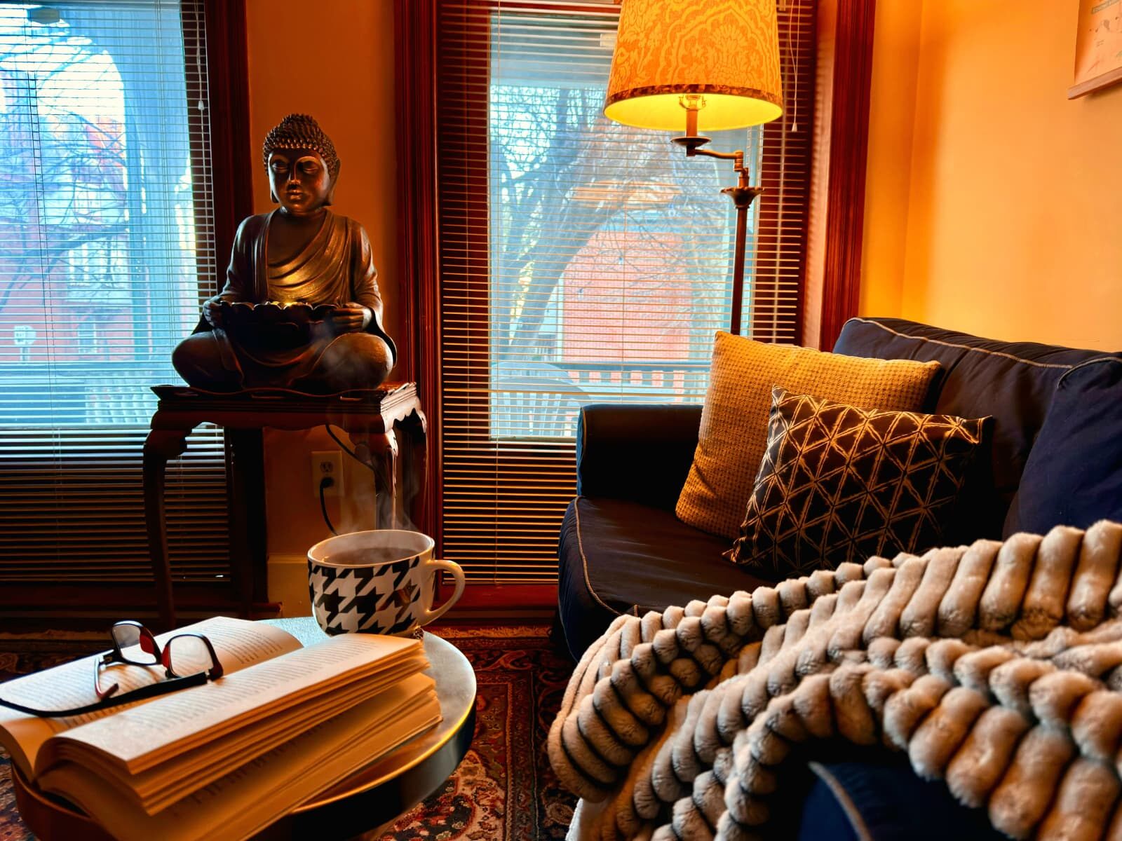 A blue sofa with a knitted throw under a warm lamp, a cup of tea steaming beside an open book.