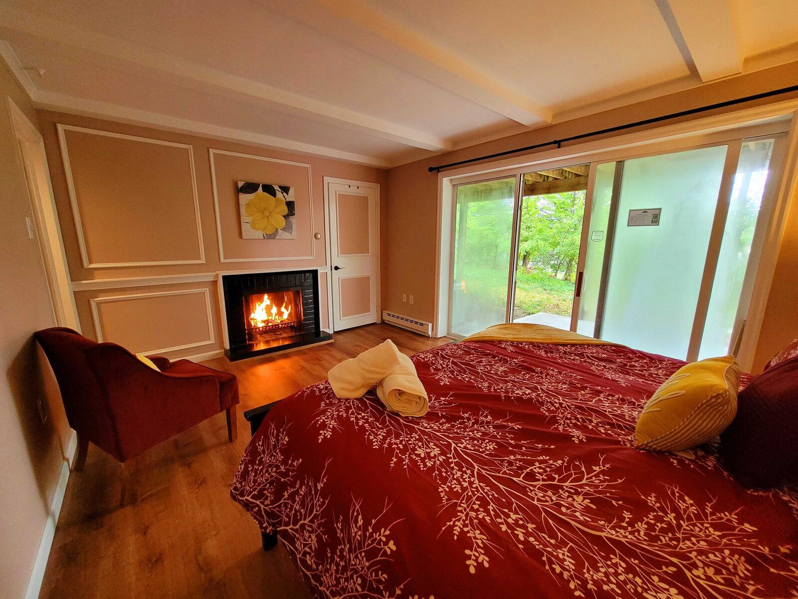 A bed made up in red with two folded towels, a fire lit in the hearth and the garden beyond the glass doors.