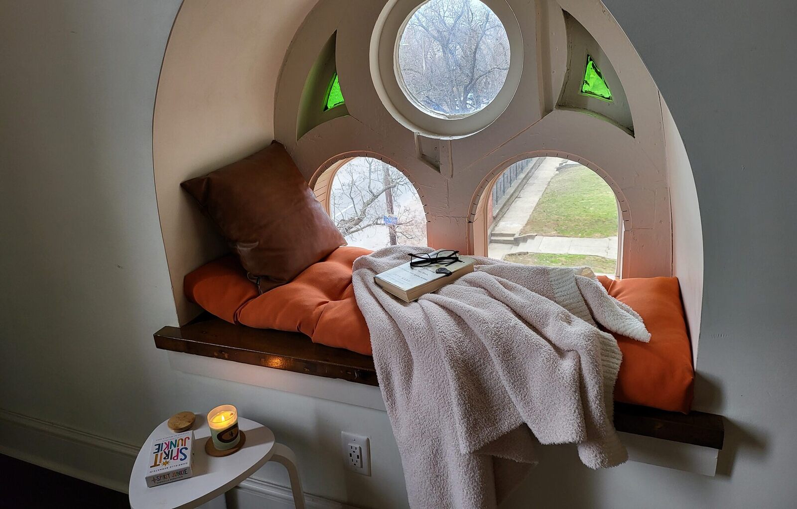 A window seat under an arched church window, a cushion and a throw, a book left open.