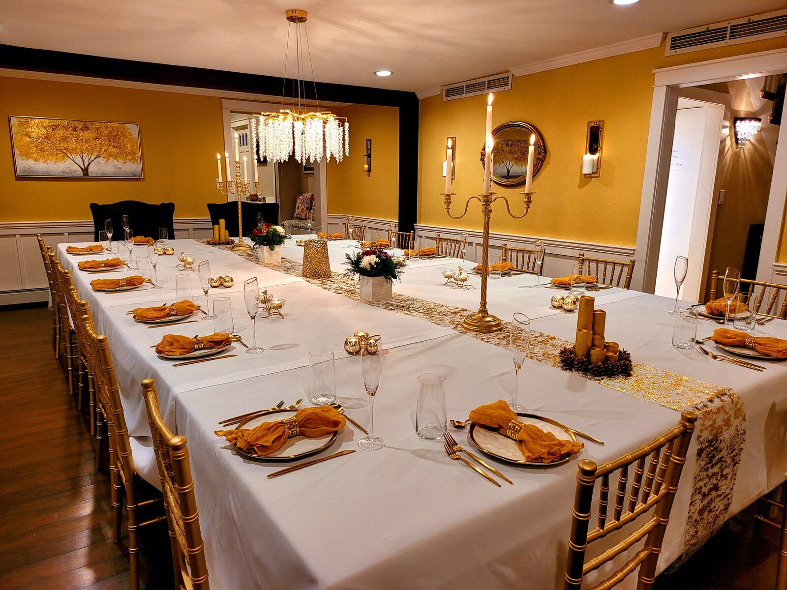 The long table in the grand dining room set for twenty, white cloth, gold candelabra and amber napkins under a crystal chandelier.